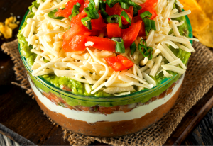 Mexican Layer Dip which is layers of mexican beans, sour cream, avocado with cheese and tomato on top