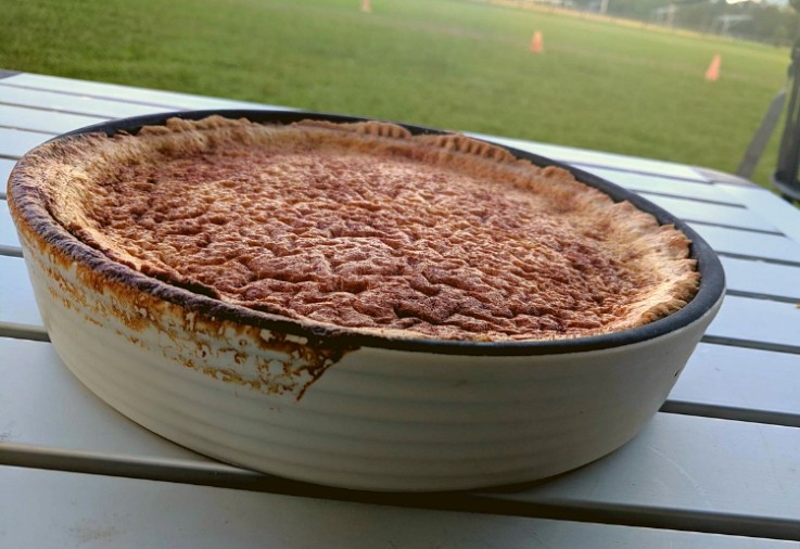 Ovenproof baking dish sitting on an outdoor table containing a delicious milk based tart with a golden top sprinkled with cinnamon