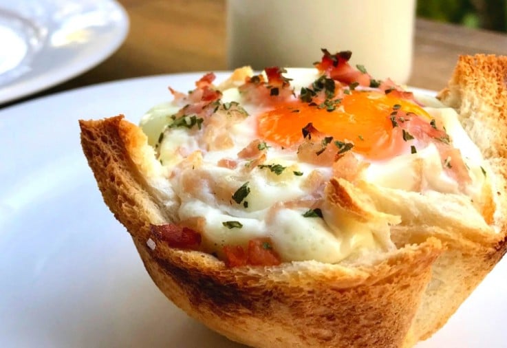 Close up picture of a tart made from a slice of toast filled with egg and bacon with parsley on top served on a white plate