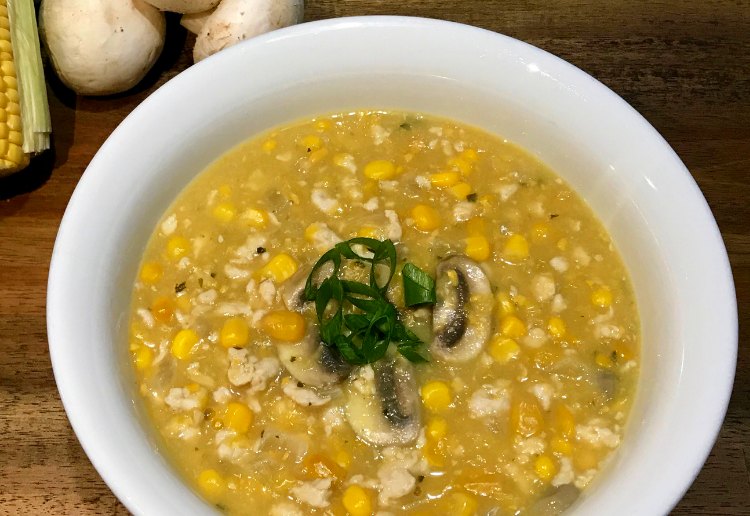 A large white bowl filled with hearty chicken and corn soup garnished with spring onions and also with mushrooms