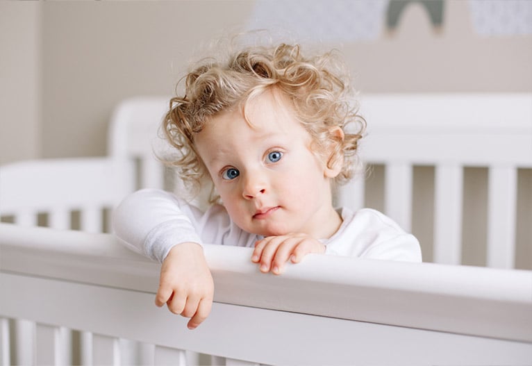 Toddler standing in cot looking at camera.