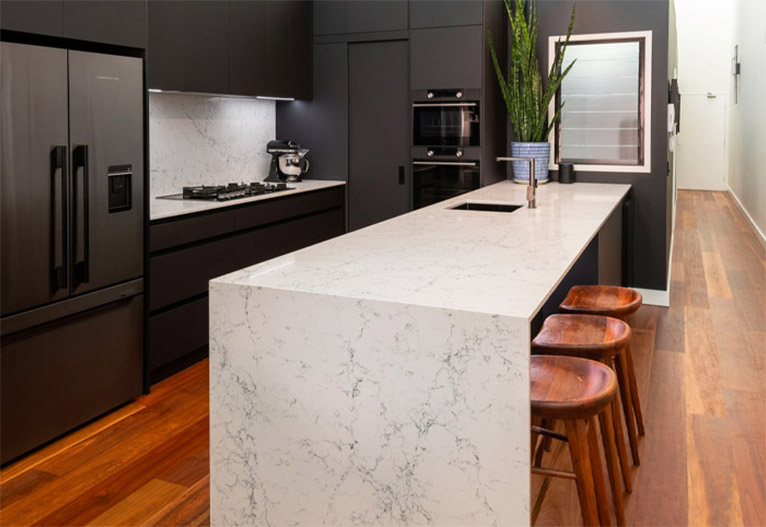 White stone waterfall benchtop in a black kitchen with wooden stools.