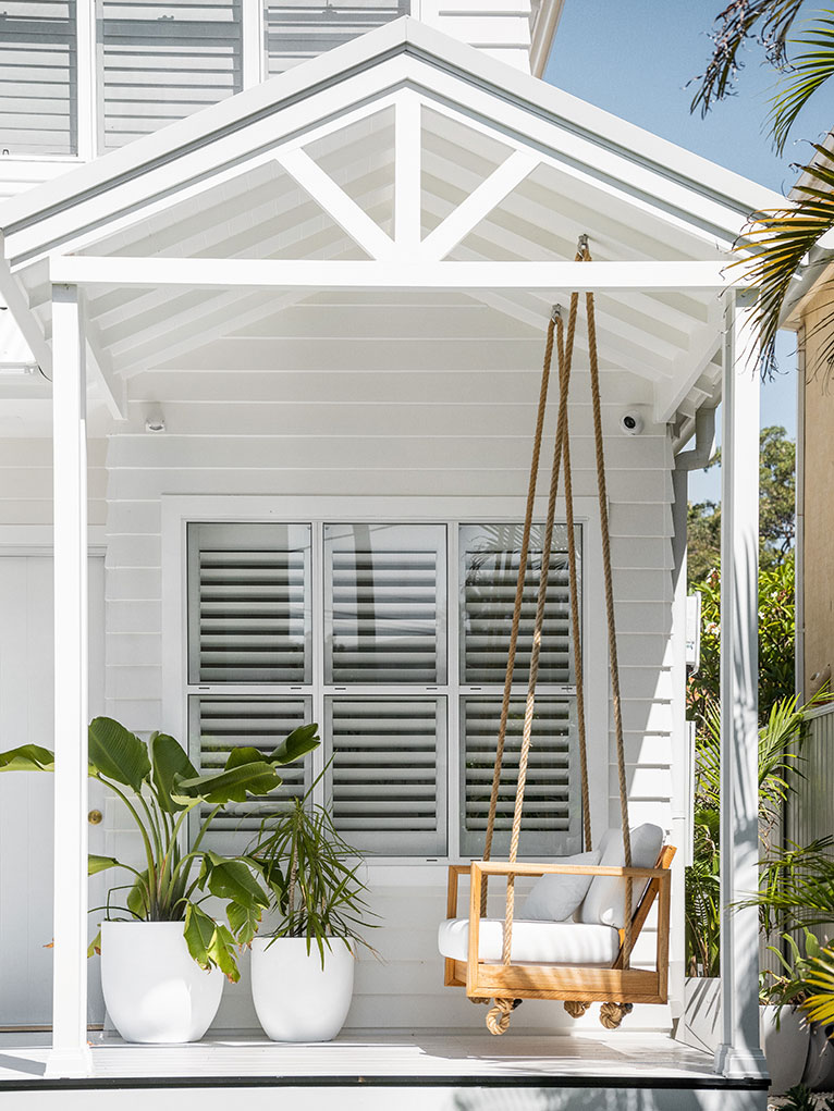 Porch of a white beach cottage with a swing seat and pot plants.