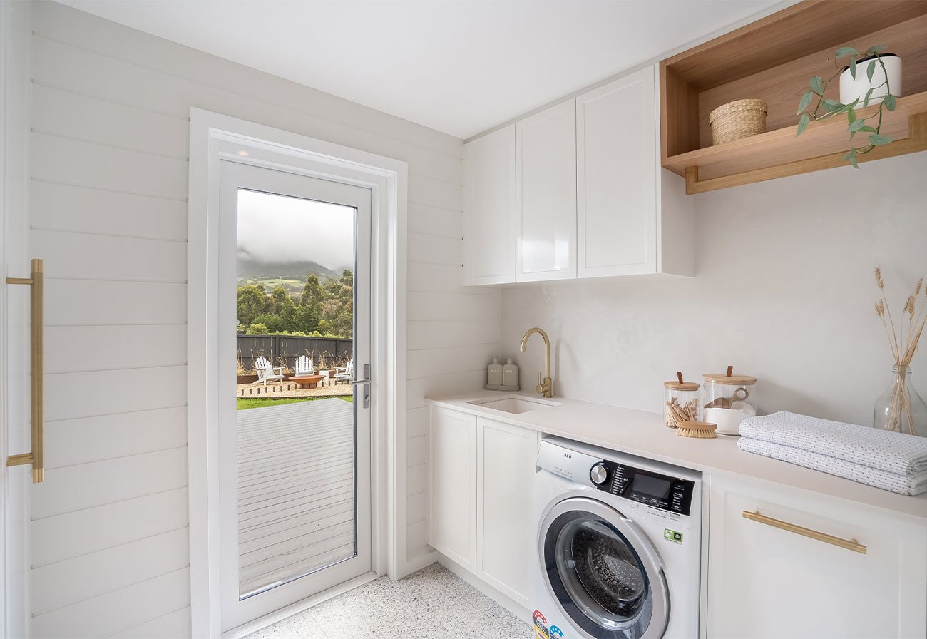 White laundry with brass tapware and glass door opening to a deck.