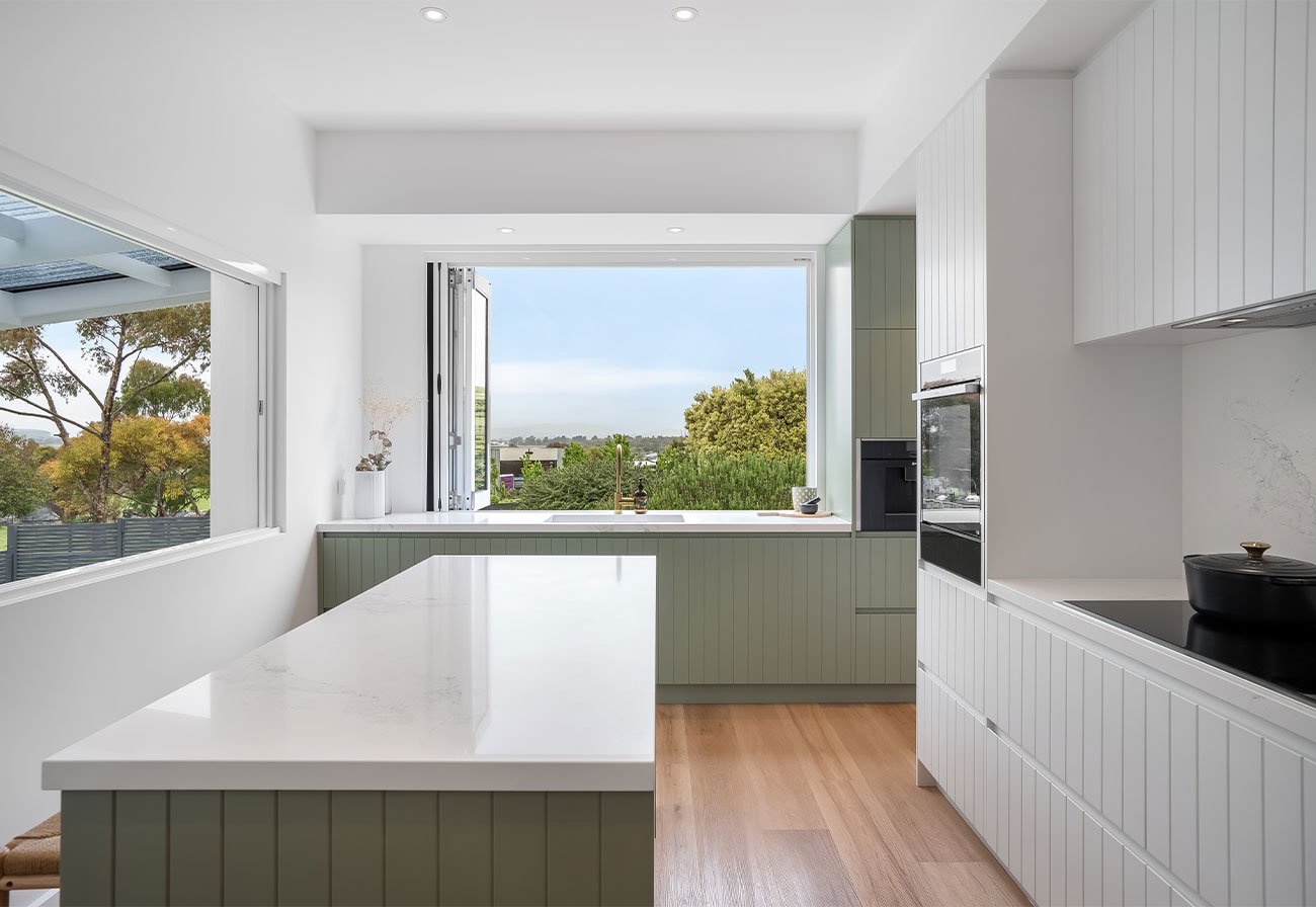 Modern kitchen with green cabinets, white benches and a window overlooking a green landscape.