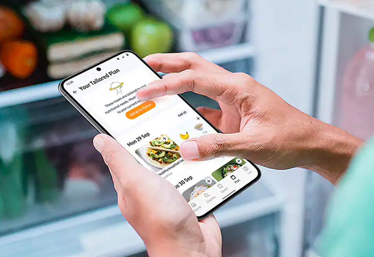 Person using a food app on a mobile phone while standing in front of an open fridge.